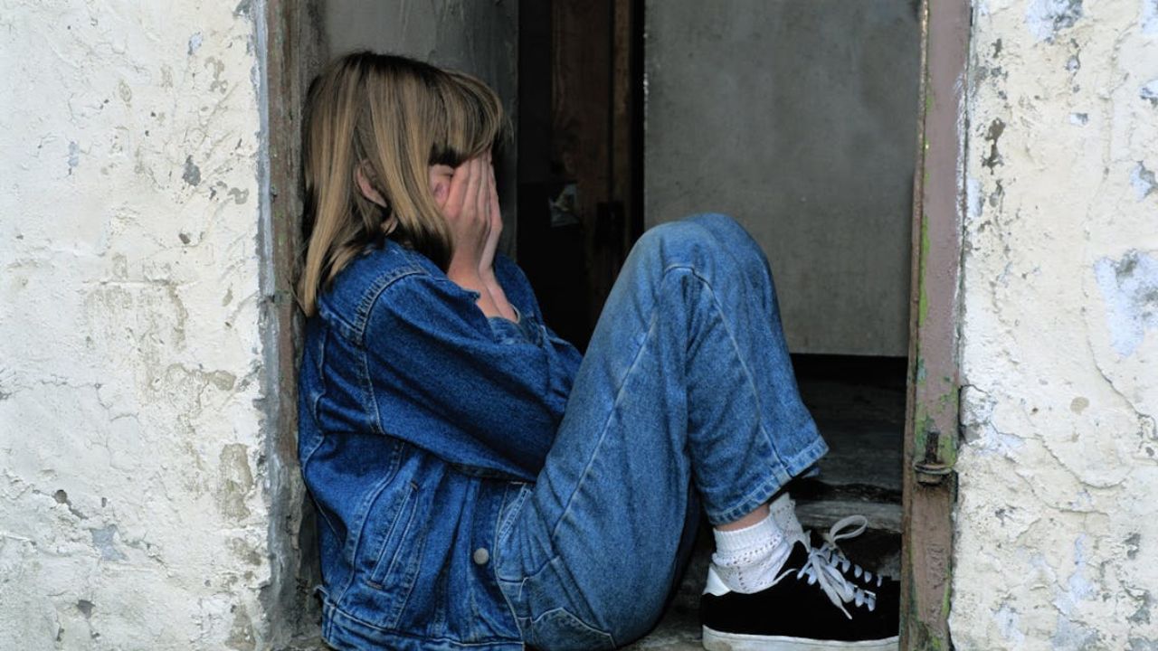 Niña víctima de alienación parental vestida con pantalón y chamarra de mezclilla azul con tenis negros sentada con las manos tapándole la cara