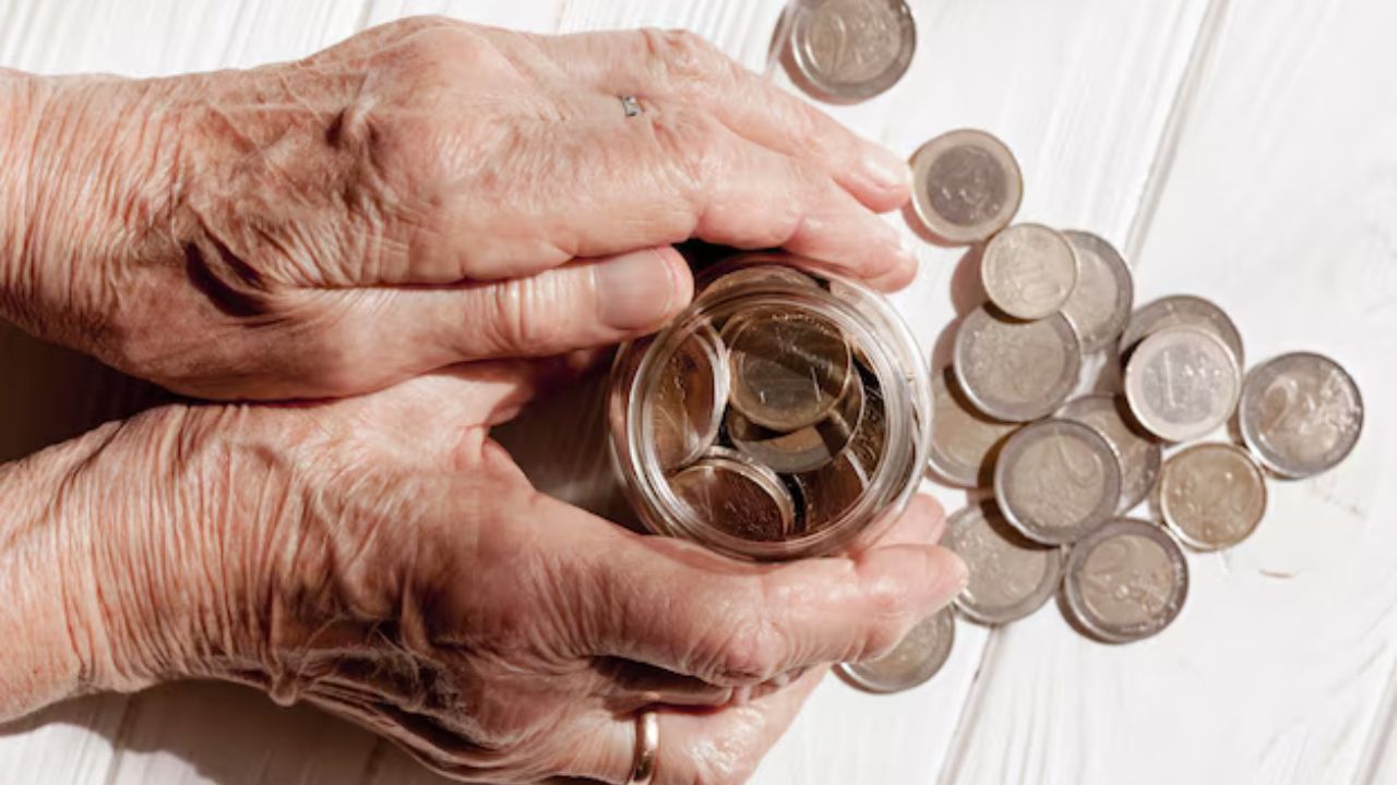 Manos de un adulto mayor sosteniendo un frasco lleno de monedas sobre una mesa donde hay monedas sueltas. 