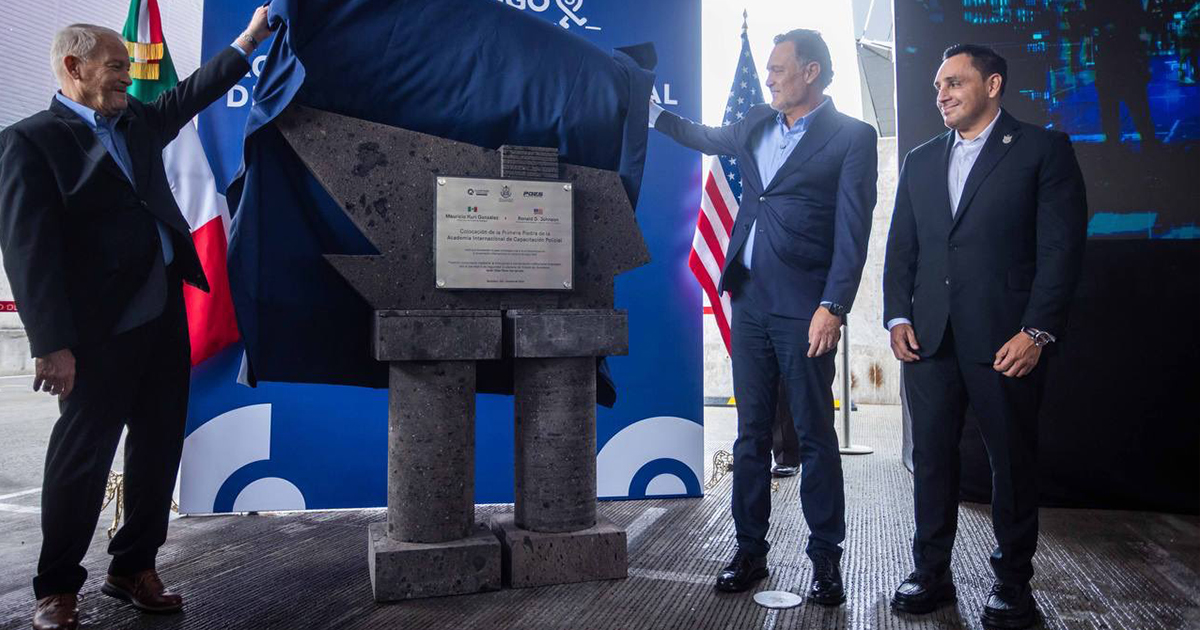 develamiento de una placa conmemorativa en un evento gubernamental e internacional. A la izquierda, un funcionario levanta una tela azul para revelar la placa de piedra; al centro, el gobernador de Querétaro, Mauricio Kuri González, sostiene el otro extremo del velo junto a la bandera de Estados Unidos; a la derecha, otro funcionario en traje oscuro observa sonriente. La placa metálica montada sobre una estructura de cantera negra con dos columnas conmemora la colocación de la primera piedra de la Academia Internacional de Capacitación Policial en Querétaro.