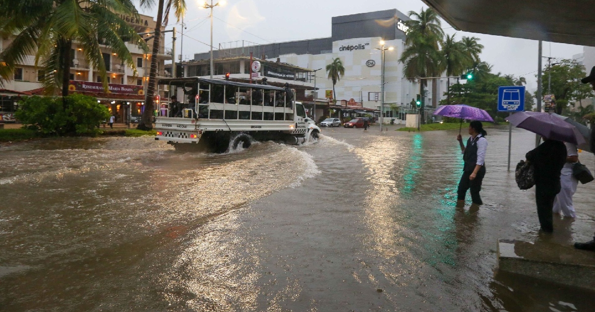 avenida costera inundada en Acapulco, Guerrero, bajo lluvia persistente. En el centro de la vialidad, un camión de la Guardia Nacional avanza generando olas a su paso por el agua acumulada. A la derecha, transeúntes con sombrillas permanecen de pie con el agua cubriéndoles los tobillos cerca de una parada de transporte público, mientras al fondo se observan palmeras, hoteles y un centro comercial (Cinépolis).