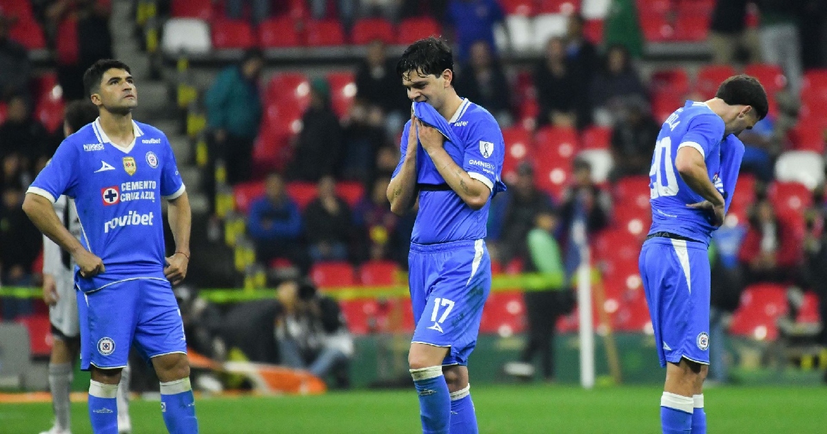 Descripción visual de la imagenPlano entero en el terreno de juego que muestra a tres futbolistas del Cruz Azul luciendo el uniforme azul de visitante/local con evidentes gestos de desilusión y frustración tras una jugada o derrota. En el centro, Amaury García (#17) se lleva el cuello de la camiseta a la boca en señal de lamento. A la izquierda, un compañero permanece de pie con las manos en las caderas mirando al horizonte, mientras que a la derecha, otro jugador con el dorsal 40 camina de espaldas con la cabeza agachada.