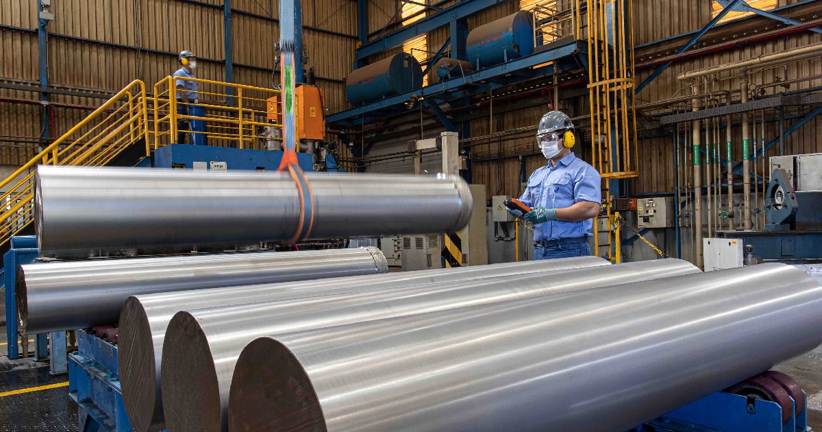 planta industrial de procesamiento metalúrgico de la empresa CBMM (Companhia Brasileira de Metalurgia e Mineração). En primer plano destacan grandes lingotes o cilindros macizos de aleación metálica de niobio con acabado pulido. Al fondo, un operario con equipo de protección personal (casco gris, lentes de seguridad, cubrebocas y protectores auditivos amarillos) opera un control remoto industrial