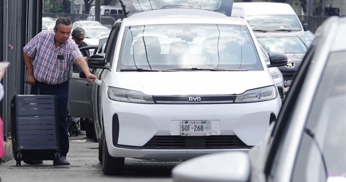 Plano medio horizontal enfocado en un automóvil eléctrico blanco de la marca china BYD, con placas de circulación de la Ciudad de México (50F-268), estacionado en la vía pública. Del lado izquierdo, un hombre maduro con camisa a cuadros azul y rosa jala una maleta negra con ruedas mientras abre la puerta trasera del vehículo para abordar o descender. Al fondo y en los laterales se aprecia tráfico vehicular continuo en una zona urbana