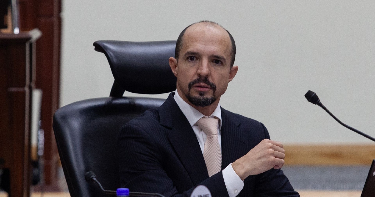 Primer plano del consejero electoral del Instituto Nacional Electoral (INE), Arturo Castillo Loza, sentado en su escaño durante una sesión del Consejo General. Viste un traje oscuro de rayas diplomáticas, camisa blanca y cravat/corbata clara, prestando atención a las intervenciones de la asamblea.