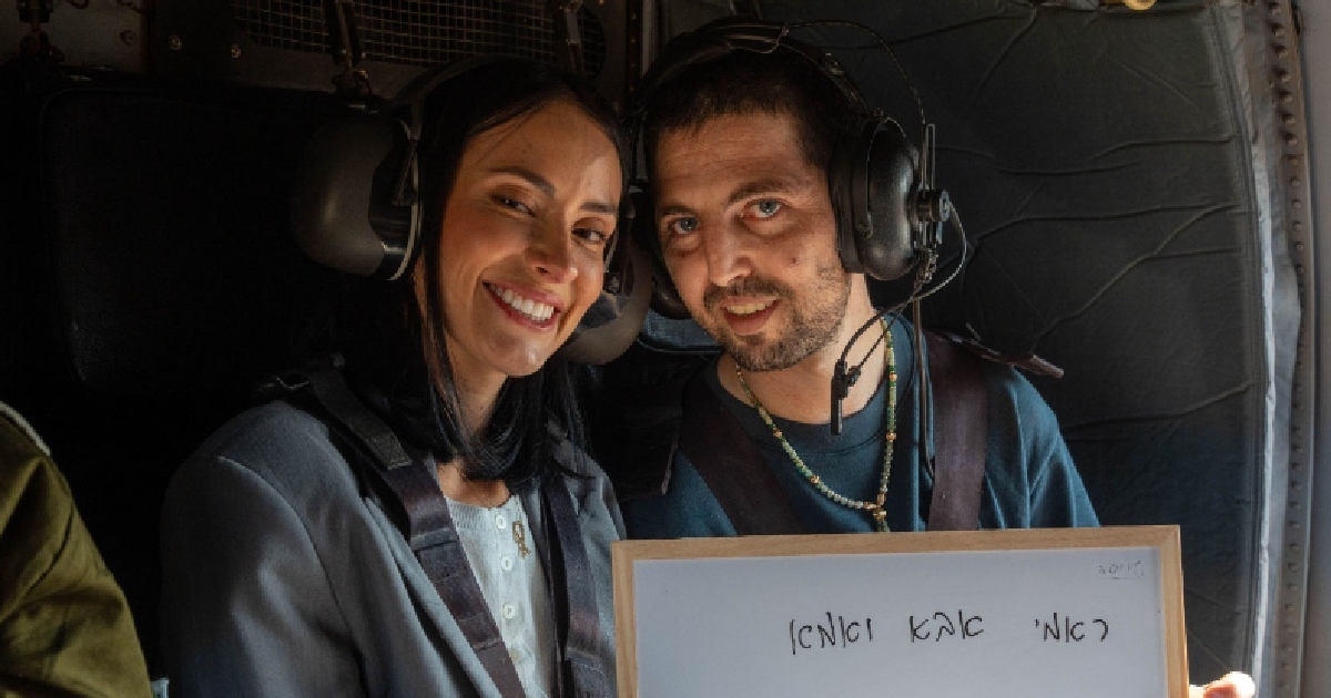 en el interior de una aeronave o helicóptero militar de transporte que muestra a Elkana Bohbot junto a su esposa Rivka. Ambos llevan audífonos de aviación con micrófono integrado y arneses de seguridad. Sonríen hacia la cámara mientras sostienen en la parte inferior un cuadro o pizarra blanca con un mensaje manuscrito en hebreo que dice «אמא ואבא אני בא» (Mamá y papá, ya voy).