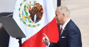 Ronald Johnson vistiendo un traje azul marino con corbata roja y pañuelo blanco en el bolsillo. Aparece levantando el índice de su mano derecha en un gesto explicativo o de énfasis mientras se desplaza frente a un atril. Al fondo destaca la Bandera Nacional de México con el Escudo Nacional en primer plano sobre la franja blanca y roja.
