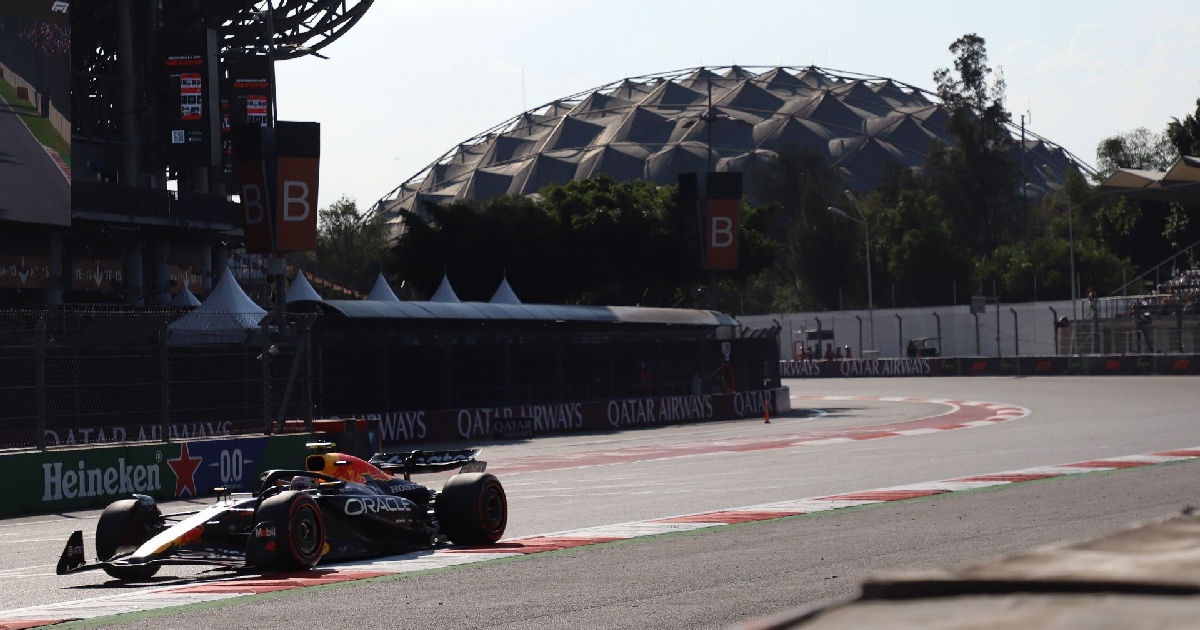 Fotografía en plano medio lateral que muestra a un monoplaza de Fórmula 1 (equipo Red Bull Racing) trazando una curva en la pista del Autódromo Hermanos Rodríguez durante el Gran Premio de México. Al fondo se aprecia la emblemática cúpula del Palacio de los Deportes rodeada de árboles y la infraestructura del circuito con vallas publicitarias de patrocinadores internacionales