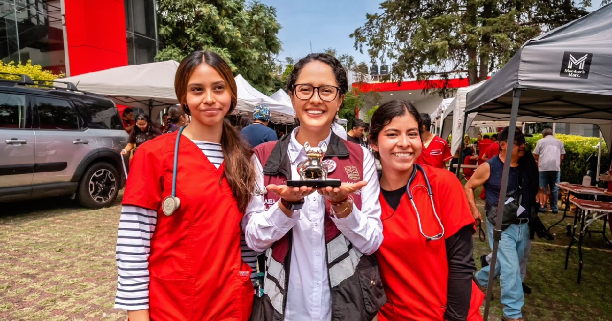 alcaldesa de Tlalpan, Gabriela Osorio (Gaby Osorio), luciendo lentes y un chaleco institucional guinda con gris de la alcaldía, sosteniendo con ambas manos una pequeña figura o reconocimiento con forma de perro. A sus lados la acompañan dos jóvenes veterinarias o personal médico de salud animal vestidas con filipinas rojas de trabajo y estetoscopios al cuello.