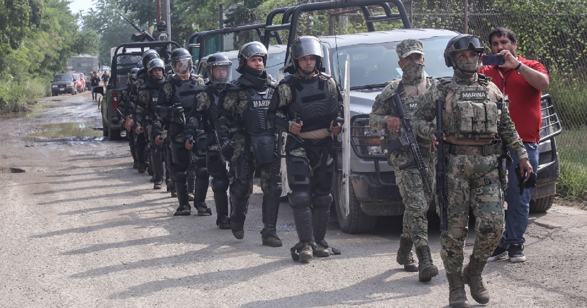 despliegue operativo de las Fuerzas Armadas y de la Marina (SEMAR) sobre un camino de terracería resguardando las inmediaciones de un centro penitenciario. A la derecha avanzan elementos militares en uniforme de camuflaje con equipo táctico y armas largas, acompañados por una fila de elementos antidisturbios portando armadura corporal completa, cascos con careta y escudos con la leyenda «MARINA». Al fondo se observan camionetas tipo pick-up de patrullaje de la institución.