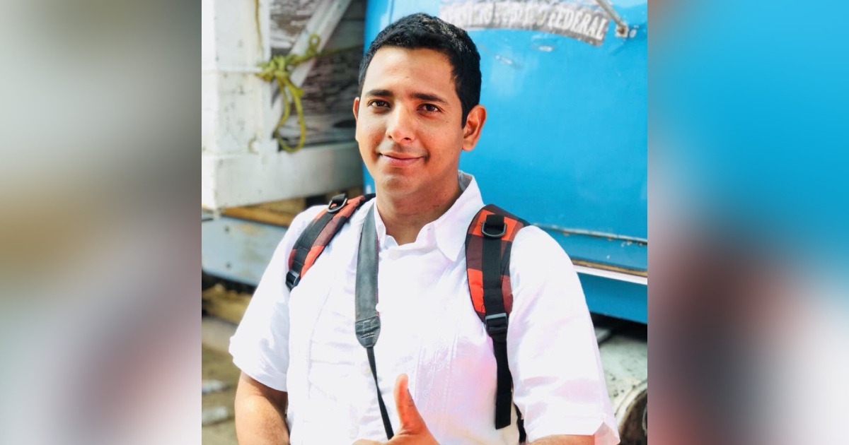 periodista Hamurabi Sinclair Huesca Manzano al aire libre. Viste una camisa/guayabera blanca de manga corta, lleva una mochila con asas a cuadros rojos y negros, y cuelga del cuello la correa negra de una cámara fotográfica. Sonríe ligeramente hacia la cámara mientras realiza el gesto de aprobación con el pulgar derecho levantado.