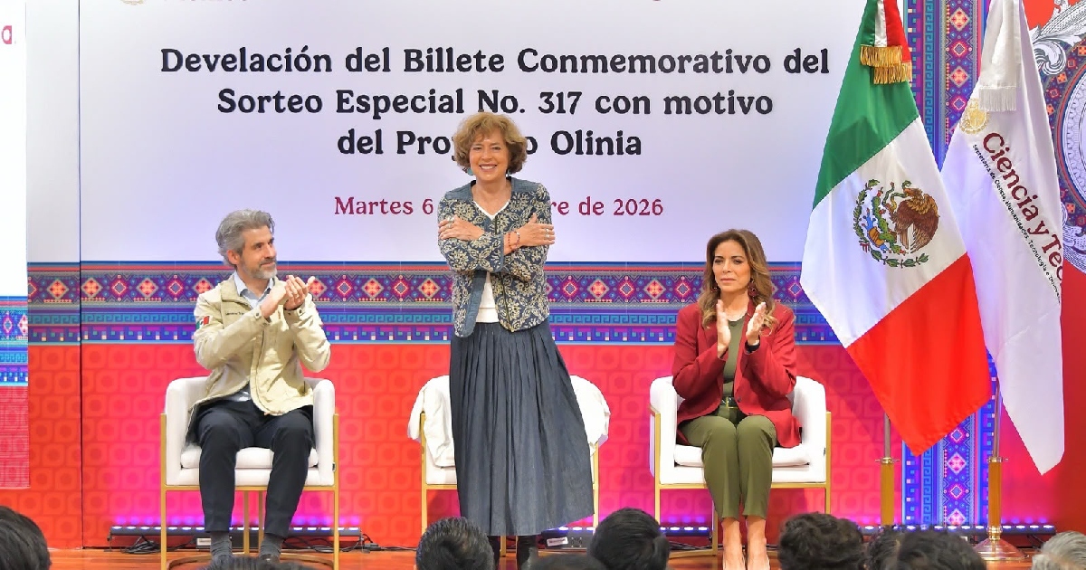 ceremonia de «Develación del Billete Conmemorativo del Sorteo Especial No. 317 con motivo del Proyecto Olinia», celebrada el martes 6 de octubre de 2026. Al centro se observa a Rosaura Ruiz Gutiérrez, titular de la Secretaría de Ciencia, Humanidades, Tecnología e Innovación, de pie con una falda larga gris y chaqueta estampada, cruzando los brazos sobre el pecho en señal de abrazo. A su izquierda (derecha de la imagen) aparece la directora general de la Lotería Nacional, Olivia Salomón Vibaldo, vistiendo un saco rojo y pantalón verde mientras aplaude sentada.