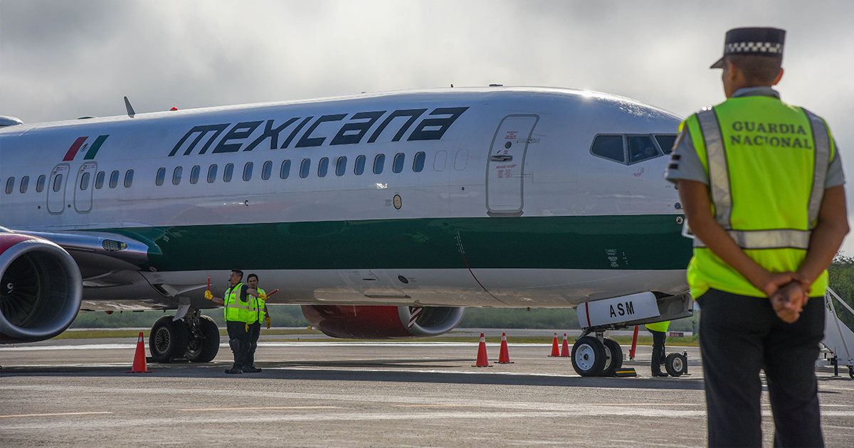 pista de un aeropuerto que muestra una aeronave comercial de la aerolínea Mexicana de Aviación. En primer plano y de espaldas, a la derecha, se encuentra un elemento de la Guardia Nacional con chaleco reflejante verde vigilando la zona operativa. En la sección media se observa el fuselaje del avión blanco con verde y la leyenda «MEXICANA» en letras oscuras, acompañado por personal de tierra con paletas de señalización dirigiendo la maniobra en la pista.