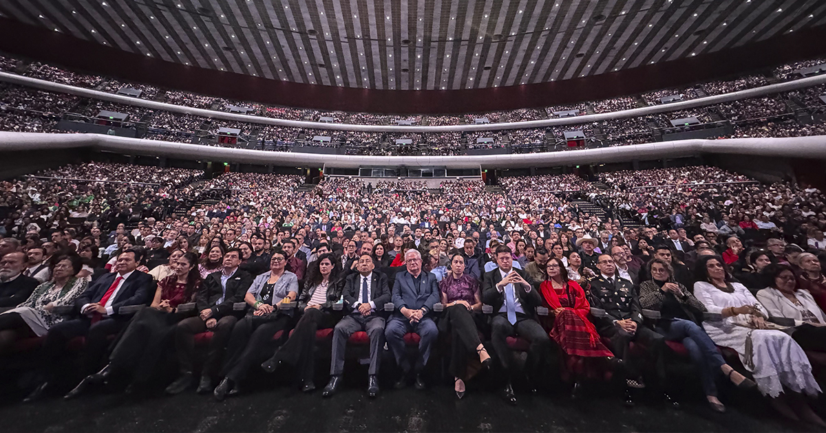Fotografía en plano general con lente gran angular desde el escenario del Auditorio Nacional de la Ciudad de México, mostrando el recinto abarrotado durante un evento del partido Morena. En la primera fila se observan funcionarios de primer nivel, legisladores e invitados especiales sentados en las butacas. Detrás de ellos, las tres secciones de gradas y balcones lucen repletas de simpatizantes bajo la imponente estructura arquitectónica y el techo de losas del reciento.
