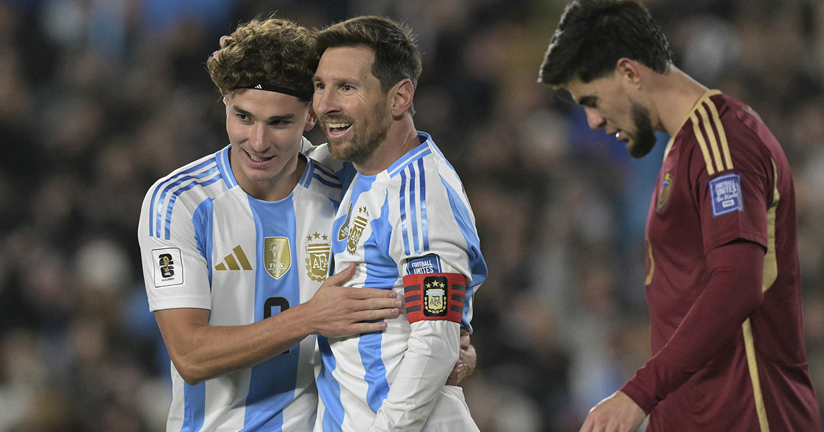 Lionel Messi y Julián Álvarez celebrando sonrientes un gol abrazados en el campo de juego con la camiseta albiceleste de la Selección Argentina. Julián Álvarez, luciendo una vincha en la cabeza, rodea a Messi por los hombros mientras este sonríe con júbilo y lleva la cinta de capitán con las tres estrellas en el brazo.