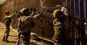 soldados de las Fuerzas Armadas de Estados Unidos (Comando Norte) instalando rollos de alambre de púas tipo concertina sobre la parte superior del muro fronterizo de acero entre México y EE. UU. Los militares visten equipo táctico completo, incluyendo uniformes camuflados, cascos con soportes de visión nocturna, chalecos balísticos y guantes tácticos. A la derecha de la estructura se aprecian marcadores numéricos dorados pintados sobre la valla metálica.