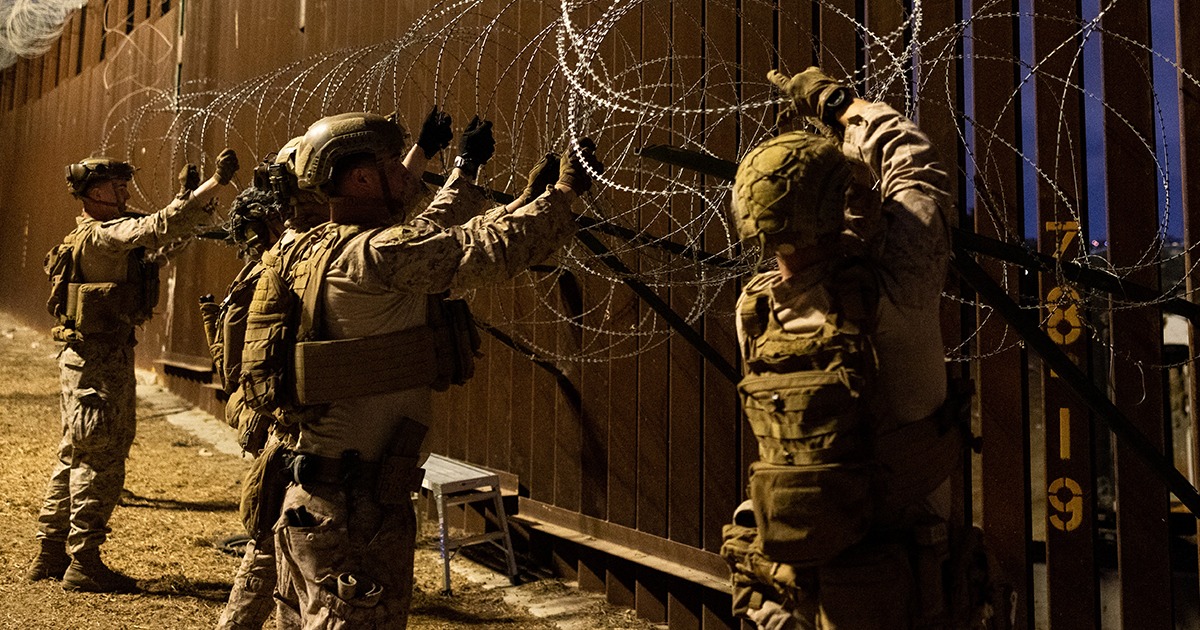 soldados de las Fuerzas Armadas de Estados Unidos (Comando Norte) instalando rollos de alambre de púas tipo concertina sobre la parte superior del muro fronterizo de acero entre México y EE. UU. Los militares visten equipo táctico completo, incluyendo uniformes camuflados, cascos con soportes de visión nocturna, chalecos balísticos y guantes tácticos. A la derecha de la estructura se aprecian marcadores numéricos dorados pintados sobre la valla metálica.