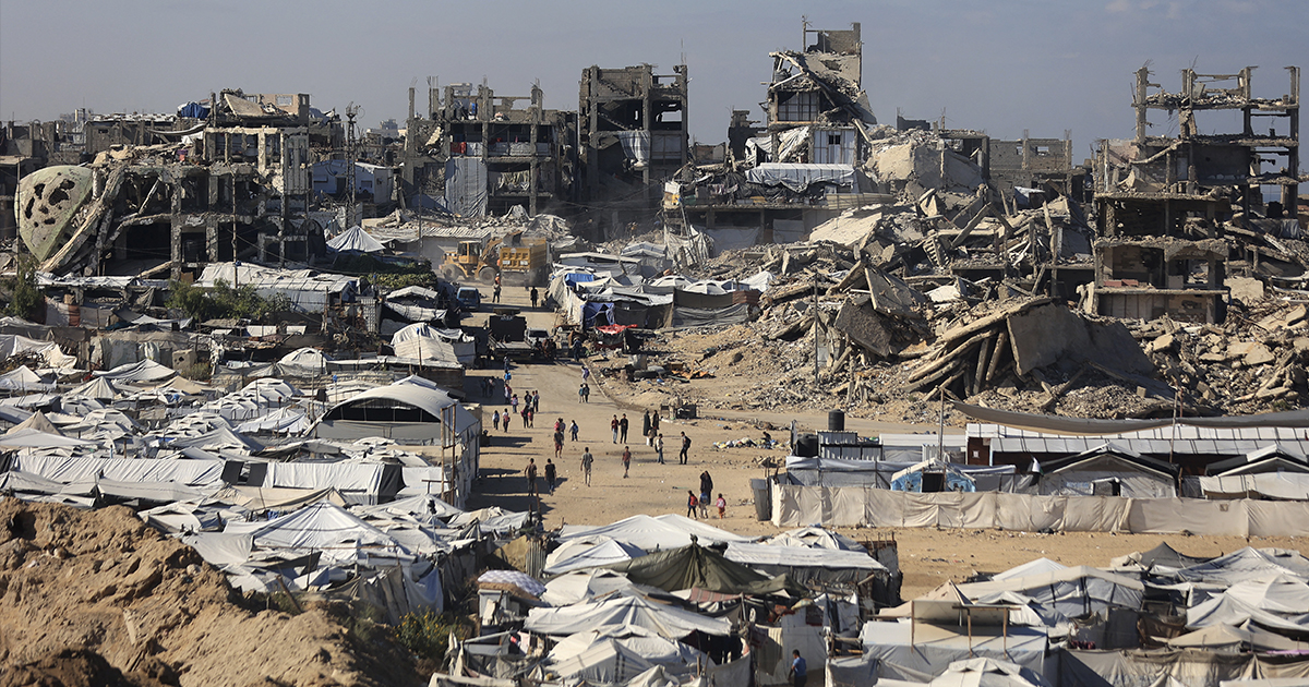devastación masiva en un paisaje urbano de la Franja de Gaza. En la mitad superior se aprecian múltiples edificios de concreto reducidos a escombros, varillas expuestas y estructuras colapsadas por los bombardeos. En la mitad inferior y primer plano se extiende un campamento improvisado repleto de carpas y tiendas de campaña blancas de refugiados, por donde transitan pequeños grupos de personas entre los caminos de tierra y ruinas.   