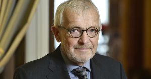 primer plano medio del físico belga Francis Halzen galardonado con el Premio Nobel de Física. El reconocido científico aparece mirando fijamente a la cámara con una ligera sonrisa, luciendo lentes de armazón circular oscuro, cabello cano, barba corta recortada y un traje formal oscuro con camisa gris y corbata a rayas.
