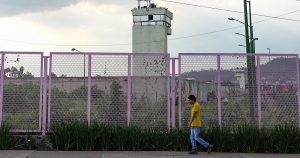 exterior de un centro penitenciario o reclusorio en la Ciudad de México. En primer plano, un joven con playera amarilla y jeans camina sobre la banqueta a lo largo de una reja o valla metálica de protección perimetral pintada de color rosa/morado. Detrás del enrejado se aprecia el muro de seguridad del penal y se erige una alta torre de vigilancia gris con ventanales superiores. Al fondo, bajo un cielo nublado, se distinguen cerros y vegetación periférica.