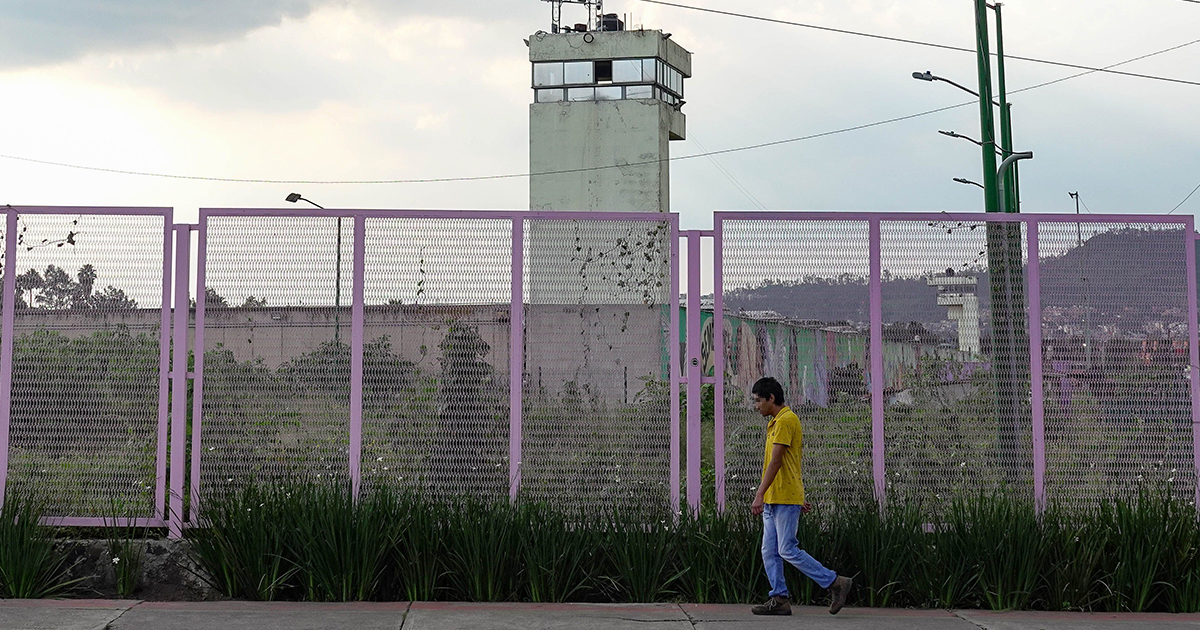 exterior de un centro penitenciario o reclusorio en la Ciudad de México. En primer plano, un joven con playera amarilla y jeans camina sobre la banqueta a lo largo de una reja o valla metálica de protección perimetral pintada de color rosa/morado. Detrás del enrejado se aprecia el muro de seguridad del penal y se erige una alta torre de vigilancia gris con ventanales superiores. Al fondo, bajo un cielo nublado, se distinguen cerros y vegetación periférica.