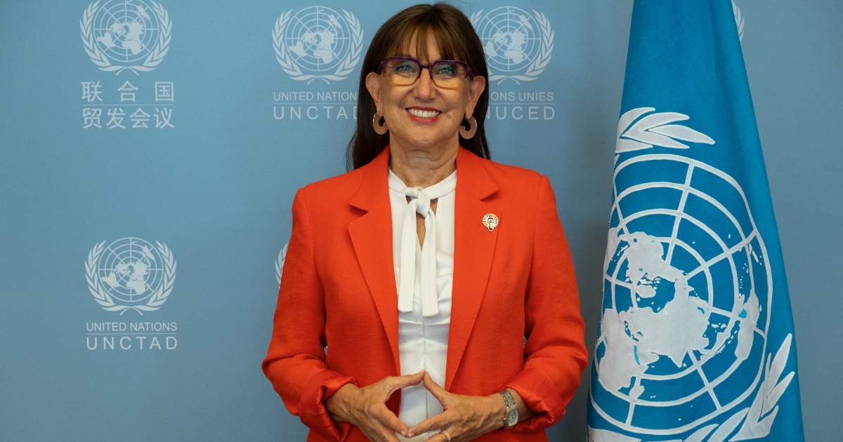 Rebeca Grynspan sonriendo de frente a la cámara. Viste un saco de color naranja/coral sobre una blusa blanca con lazada al cuello y lleva gafas de armazón oscuro.