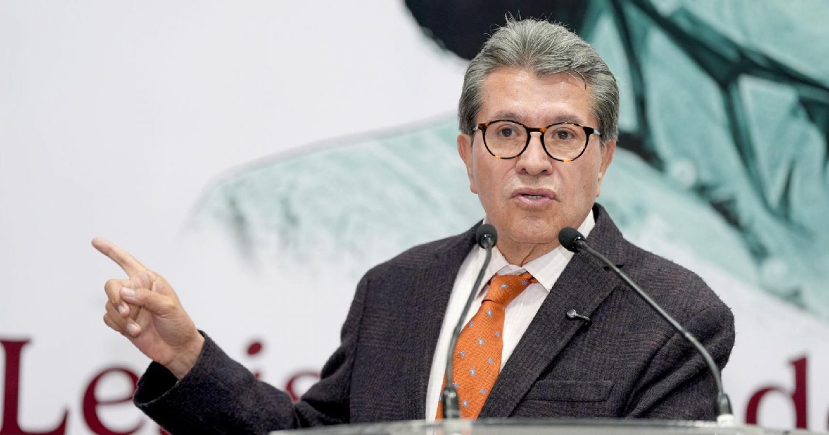 Ricardo Monreal Ávila en un podio con micrófonos, durante una conferencia de prensa o intervención pública. Viste un saco oscuro sobre camisa clara con una corbata en tono anaranjado o cobrizo, lleva gafas con armazón de carey y extiende el brazo y dedo índice derecho hacia un costado mientras se expresa con gesto enfático.