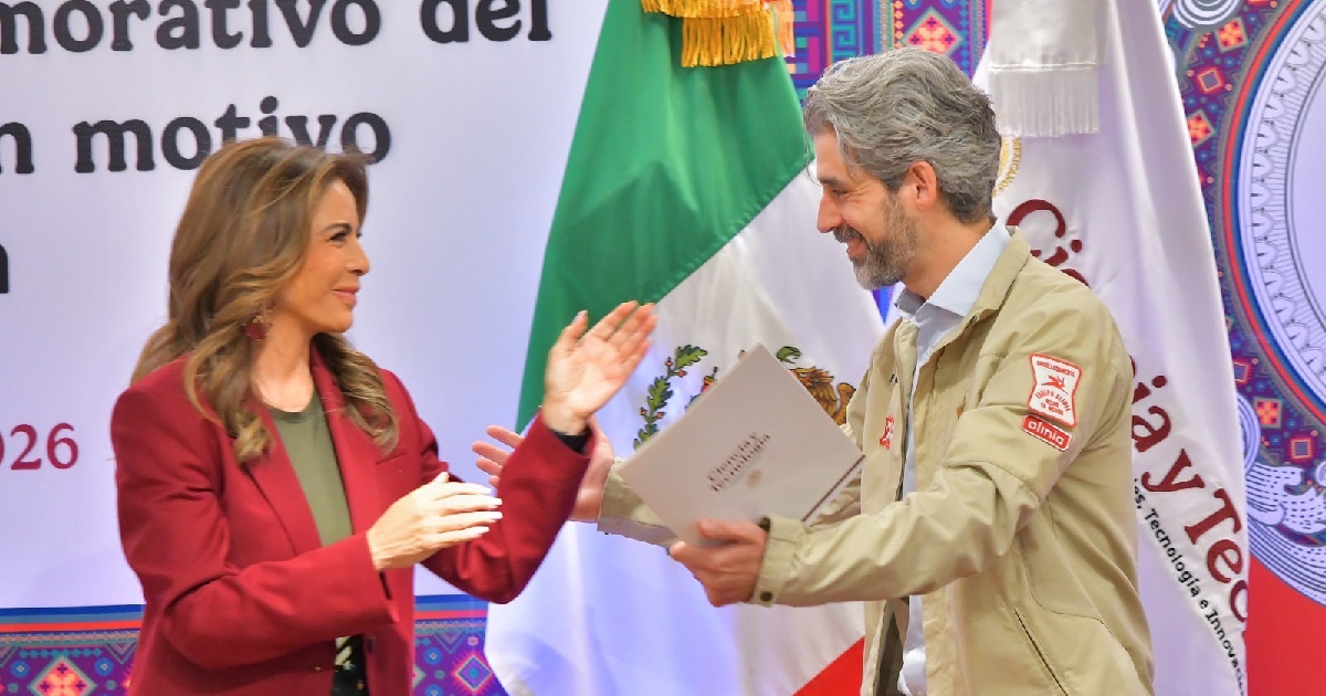 Lotería Nacional, Olivia Salomón Vibaldo, con saco color rojo borgoña, haciendo entrega formal de una carpeta o reconocimiento a Roberto Capuano Tripp, encargado del proyecto del auto eléctrico mexicano «Olinia».