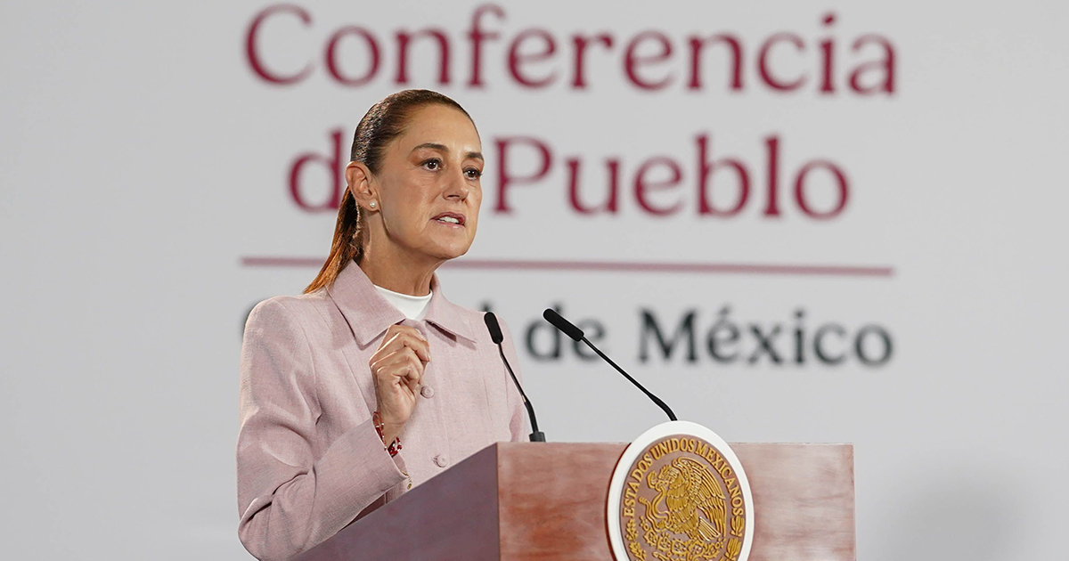 Claudia Sheinbaum Pardo, dirigiéndose al público desde el podio oficial de Palacio Nacional. La mandataria viste un saco estilizado de tono rosa pastel sobre una blusa blanca y lleva el cabello recogido en una coleta. Gesticula con la mano derecha a la altura del pecho mientras habla frente a dos micrófonos. Al fondo se distingue la mampara blanca con las letras guinda del lema institucional «Conferencia del Pueblo» y «Gobierno de México», mientras que el podio luce al frente el escudo de los Estados Unidos Mexicanos grabado en dorado sobre madera.
