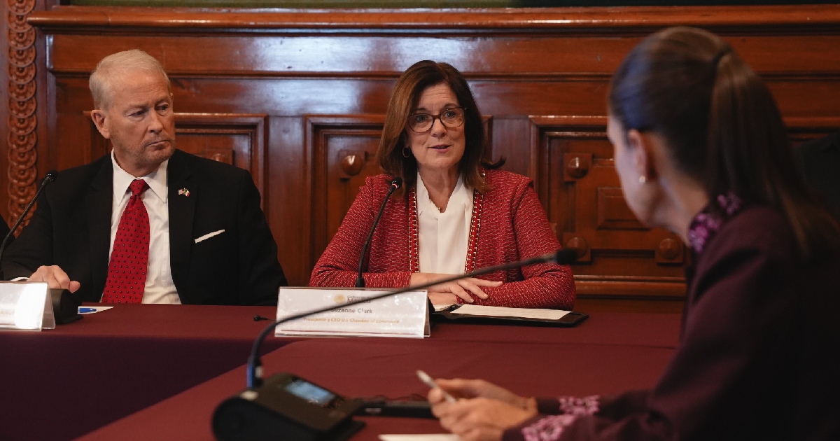 En el primer plano, a la derecha y de espaldas en escorzo, se encuentra la presidenta Claudia Sheinbaum Pardo tomando notas con un bolígrafo. Frente a ella se observan Suzanne Clark, presidenta y CEO de la U.S. Chamber of Commerce, vistiendo un saco rojo de textura tejida y blusa blanca mientras se dirige a la mandataria; a su lado izquierdo está sentado el diplomático estadounidense Ronald Johnson con traje oscuro, camisa blanca y corbata roja con el pin de las banderas de México y EE. UU. en la solapa. Sobre la mesa destacan micrófonos e identificadores oficiales.