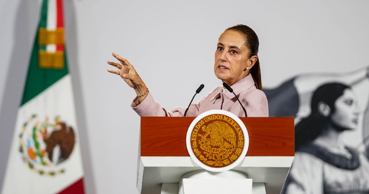 Claudia Sheinbaum Pardo, dirigiéndose a la audiencia desde el podio oficial en Palacio Nacional. Porta un saco rosa claro y extiende la mano derecha abierta hacia adelante en un gesto explicativo durante su discurso. A la izquierda de la toma se encuentra la Bandera Nacional mexicana y a la derecha una ilustración de heroínas de la historia patria. Al frente, el podio exhibe el escudo oficial de los Estados Unidos Mexicanos sobre acabado de madera.