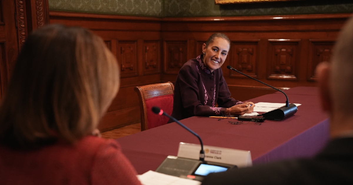 Claudia Sheinbaum Pardo, presidiendo una mesa de trabajo en un salón con acabados en madera de Palacio Nacional. La mandataria viste un saco de color berenjena/morado con bordados artesanales en puños y cuello, y se muestra sonriente conversando hacia sus interlocutores. En el primer plano, desenfocados en escorzo, se aprecia la silueta de Suzanne Clark (izquierda, vestida de rojo) y la del embajador de Estados Unidos (derecha), con micrófonos e identificadores oficiales sobre el mantel tinto.
