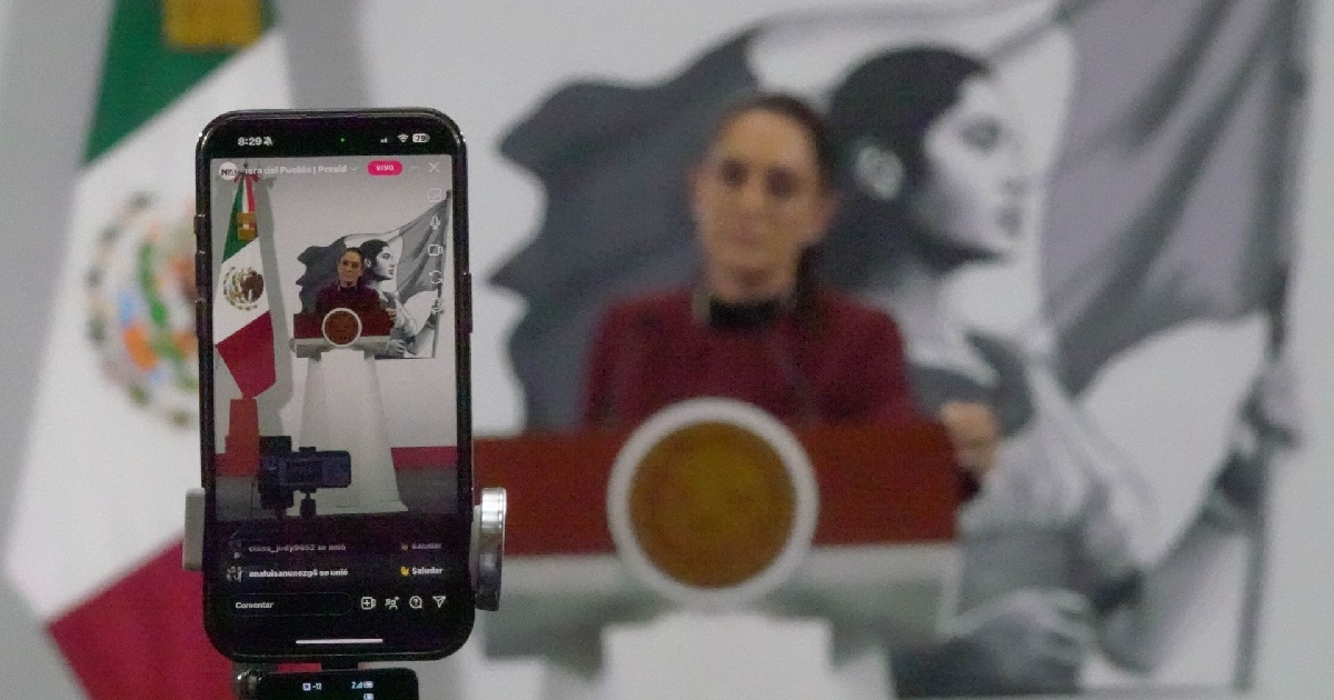 Plano detalle en profundidad de campo reducida que enfoca la pantalla de un teléfono inteligente (smartphone) colocado sobre un trípode transmitiendo en vivo en redes sociales. En la pantalla del dispositivo se aprecia la imagen vertical de la presidenta Claudia Sheinbaum pardo en el podio durante la conferencia «La Mañanera del Pueblo», con la bandera de México a la izquierda, comentarios de los usuarios e indicadores de emisión en vivo. Al fondo, de manera desenfocada, se observa a la mandataria en el estrado con el escudo nacional y el mural representativo.
