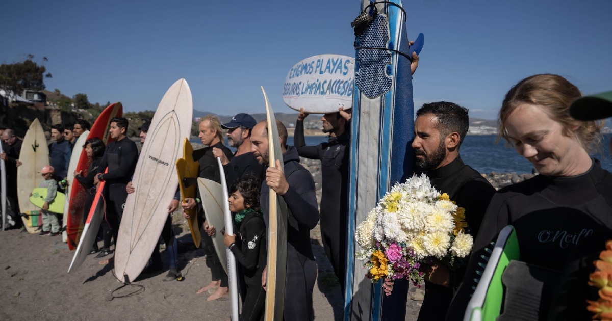 Plano medio horizontal que muestra a un grupo numeroso de surfistas reunidos en la orilla de una playa durante una protesta pacífica y homenaje memorial. Los asistentes, vestidos con trajes de neopreno (wetsuits), sostienen sus tablas de surf de manera vertical. En primer plano, a la derecha, un hombre lleva un ramo de flores blancas, amarillas y rosas junto a una tabla azul, mientras que al fondo otro manifestante levanta una tabla con el mensaje pintado: «EXIGIMOS PLAYAS SEGURAS, LIMPIAS, ACCESIBLES».