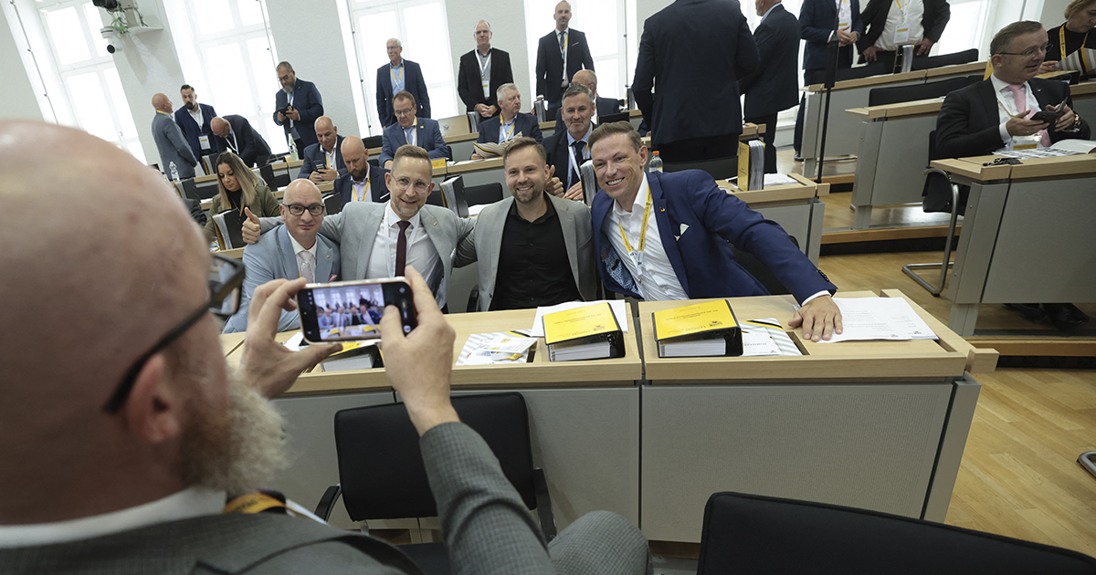 un grupo de cuatro políticos del partido de ultraderecha Alternativa para Alemania (AfD) posando sonrientes y abrazados tras sus escaños parlamentarios. En primer plano, a la izquierda, se observa la nuca y las manos de un hombre con barba que los fotografía con un teléfono inteligente. Sobre los escritorios de madera del parlamento regional se aprecian carpetas amarillas y documentos de trabajo, mientras al fondo otros miembros y asistentes caminan por el recinto.