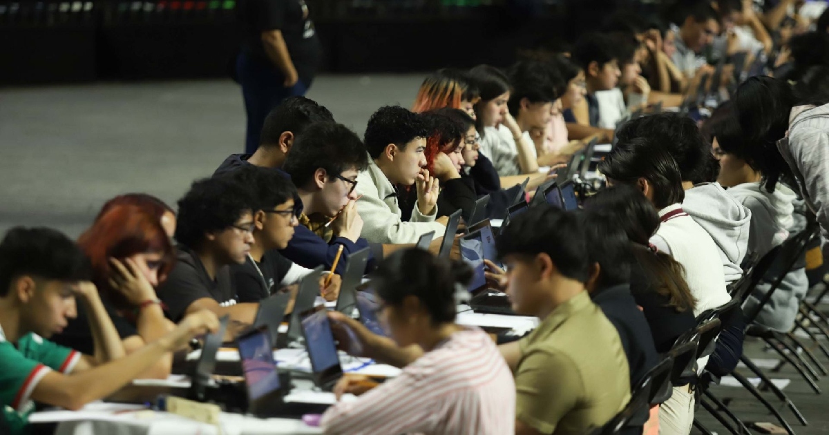 decenas de jóvenes aspirantes están sentados en largas filas alineadas. Cada estudiante trabaja concentrado frente a una computadora portátil individual colocada sobre tablones de trabajo. La iluminación del espacio está enfocada en los escritorios, dejando el fondo en un tono tenue
