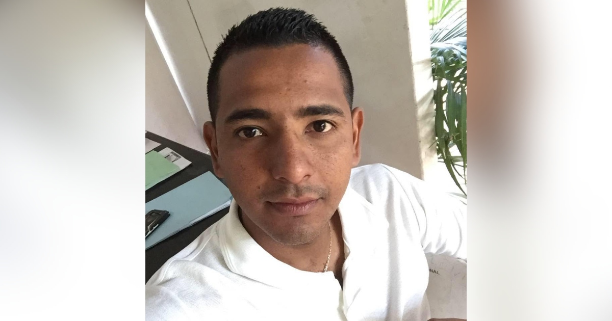 Retrato en primer plano (selfie) del periodista Hamurabi Huesca Manzano. Viste una playera polo blanca y una cadena delgada en el cuello. Muestra una expresión serena mirando directamente a la cámara. Se encuentra en un espacio de oficina con un escritorio a la izquierda, donde se aprecian carpetas o documentos en color verde y azul junto a un teléfono móvil, y vegetación al fondo a la derecha.
