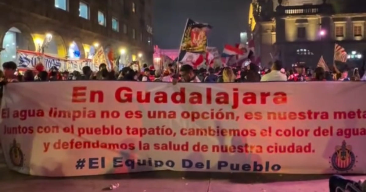 concentración de aficionados del Club Deportivo Guadalajara (Chivas) en una plaza pública urbana (área del Centro Histórico de Guadalajara). Los barristas e hinchas sostienen en primer plano una manta horizontal blanca con letras rojas y azules donde se lee: «En Guadalajara El agua limpia no es una opción, es nuestra meta. Juntos con el pueblo tapatío, cambiemos el color del agua y defendamos la salud de nuestra ciudad. #ElEquipoDelPueblo».