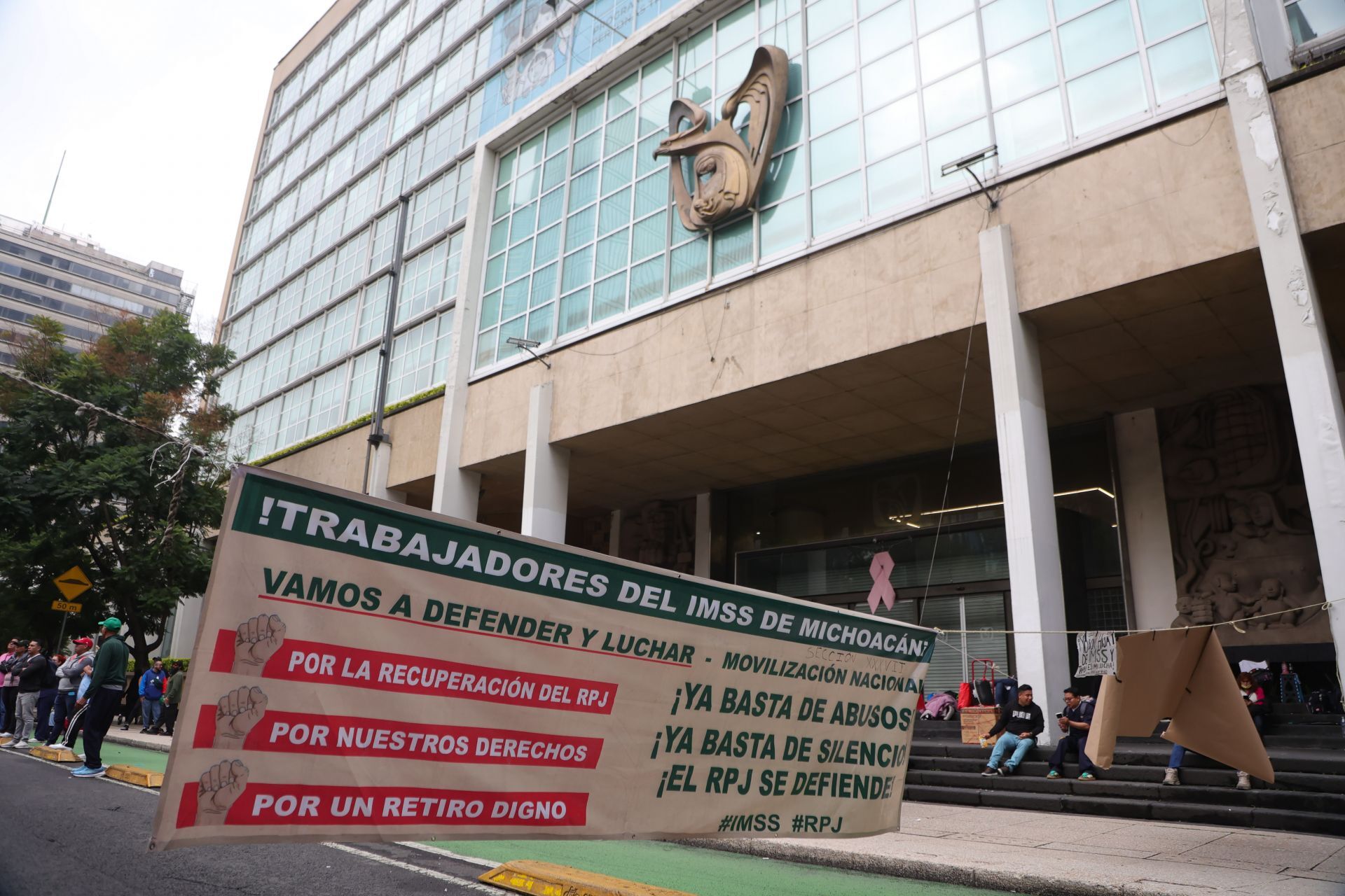 achada principal del edificio de oficinas centrales del Instituto Mexicano del Seguro Social (IMSS) sobre Paseo de la Reforma en la Ciudad de México. En el acceso exterior, frente a las escalinatas del inmueble, se observa suspendida una gran manta de protesta de la sección de Michoacán. En el manifiesto impreso en verde y rojo se lee: «!TRABAJADORES DEL IMSS DE MICHOACÁN VAMOS A DEFENDER Y LUCHAR - MOVILIZACIÓN NACIONAL ¡YA BASTA DE ABUSOS! ¡YA BASTA DE SILENCIO!