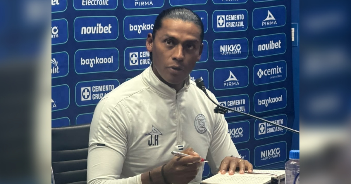 Plano medio frontal de Joel Huiqui, exjugador e integrante del cuerpo técnico del Club de Fútbol Cruz Azul, en una conferencia de prensa frente a micrófonos institucionales. Viste una sudadera de entrenamiento blanca con cierre medio de la marca Pirma, con el escudo del club en el pecho izquierdo y sus iniciales «J.H» estampadas en el derecho. Sostiene una pluma con la mano derecha mientras revisa un cuaderno de notas sobre la mesa. Al fondo se aprecia el muro de patrocinadores oficiales del equipo con los logotipos de Cemento Cruz Azul, Pirma, Novibet y Nikko.