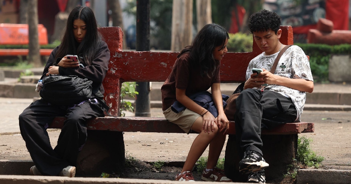 tres jóvenes adolescentes sentados en una banca de concreto en un parque público. A la izquierda, una joven vestida con sudadera y pantalones oscuros revisa atentamente la pantalla de su teléfono móvil. A la derecha, otra joven y un joven de cabello rizado conversan mientras observan juntos el dispositivo móvil que sostiene él en las manos.