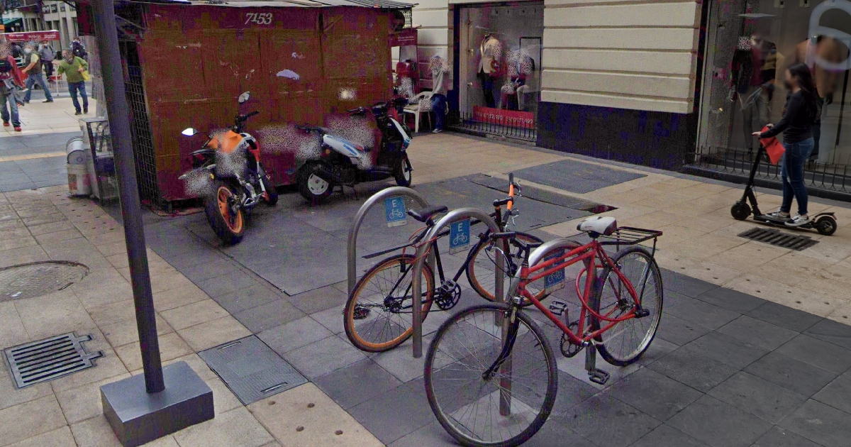 Vista de una banqueta y zona peatonal en una calle del Centro Histórico de la Ciudad de México. En el centro destacan dos bicicletas sujetas a estructuras metálicas de estacionamiento (biciestacionamiento urbano). A la izquierda, junto a un puesto metálico de periódico de color granate, se observan dos motocicletas estacionadas irregularmente sobre la acera. A la derecha, una persona transita sobre la banqueta a bordo de un monopatín o scooter eléctrico, mientras al fondo caminan otros peatones frente a los comercios.