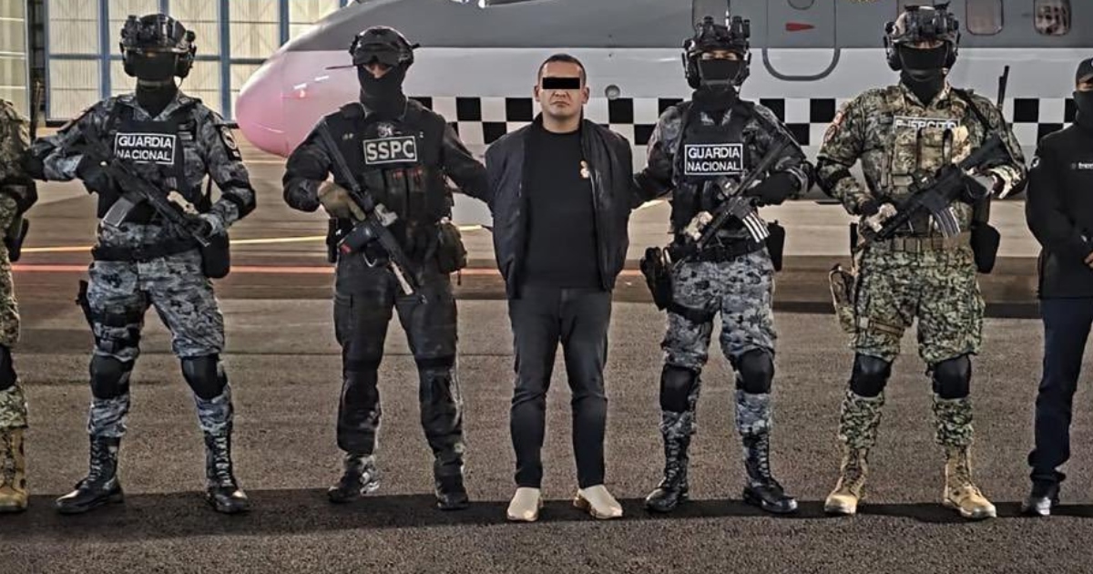 Descripción visual de la imagen Fotografía oficial de presentación en plano general que muestra al centro a un presunto operador criminal con el rostro censurado mediante una franja negra. El individuo está escoltado de pie en la pista de un aeródromo frente a una aeronave por un contingente de fuerzas de seguridad armadas y encapuchadas: dos elementos de la Guardia Nacional (GN) con uniformes camuflados grises, un agente de la Secretaría de Seguridad y Protección Ciudadana (SSPC) con uniforme táctico negro y un militar del Ejército Mexicano