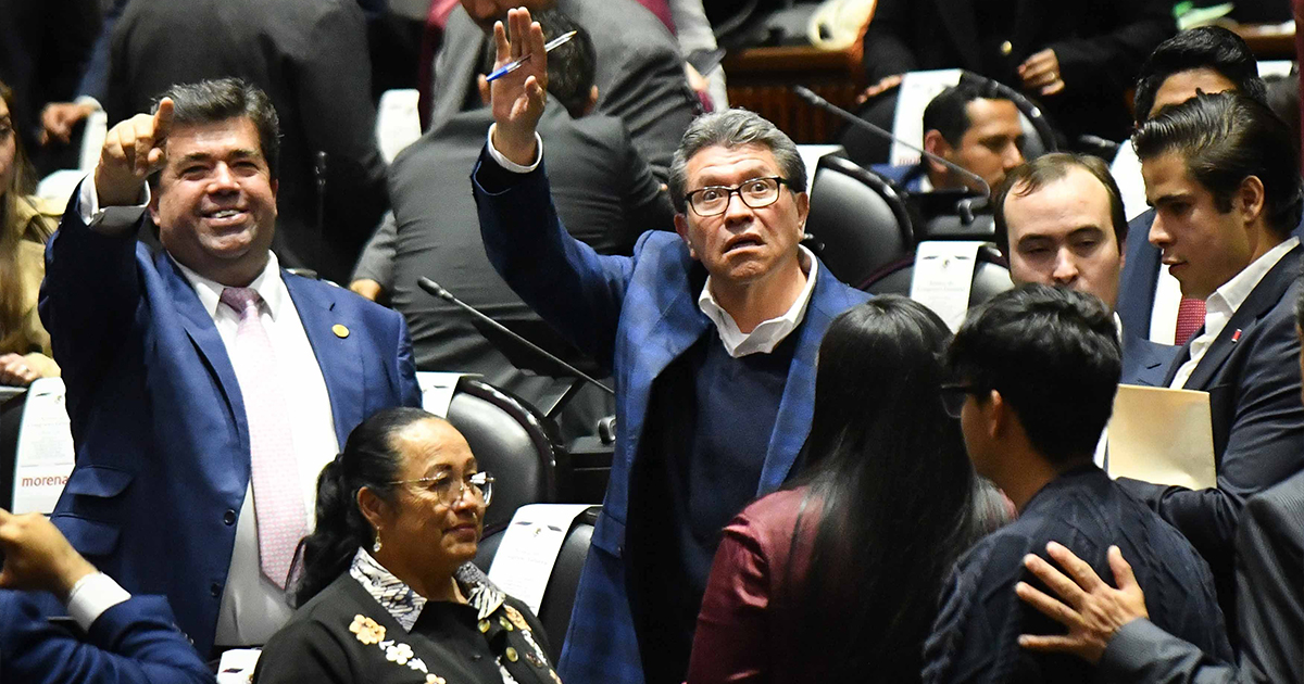 pleno de la Cámara de Diputados de México. En el centro destaca el diputado Ricardo Monreal Ávila vistiendo un saco azul marino sobre suéter del mismo tono, alzando la mano derecha con una pluma mientras observa fijamente hacia la Mesa Directiva o tribuna. A su izquierda, el legislador Pedro Haces Barba viste traje azul con corbata rosa y señala con el dedo índice hacia arriba.