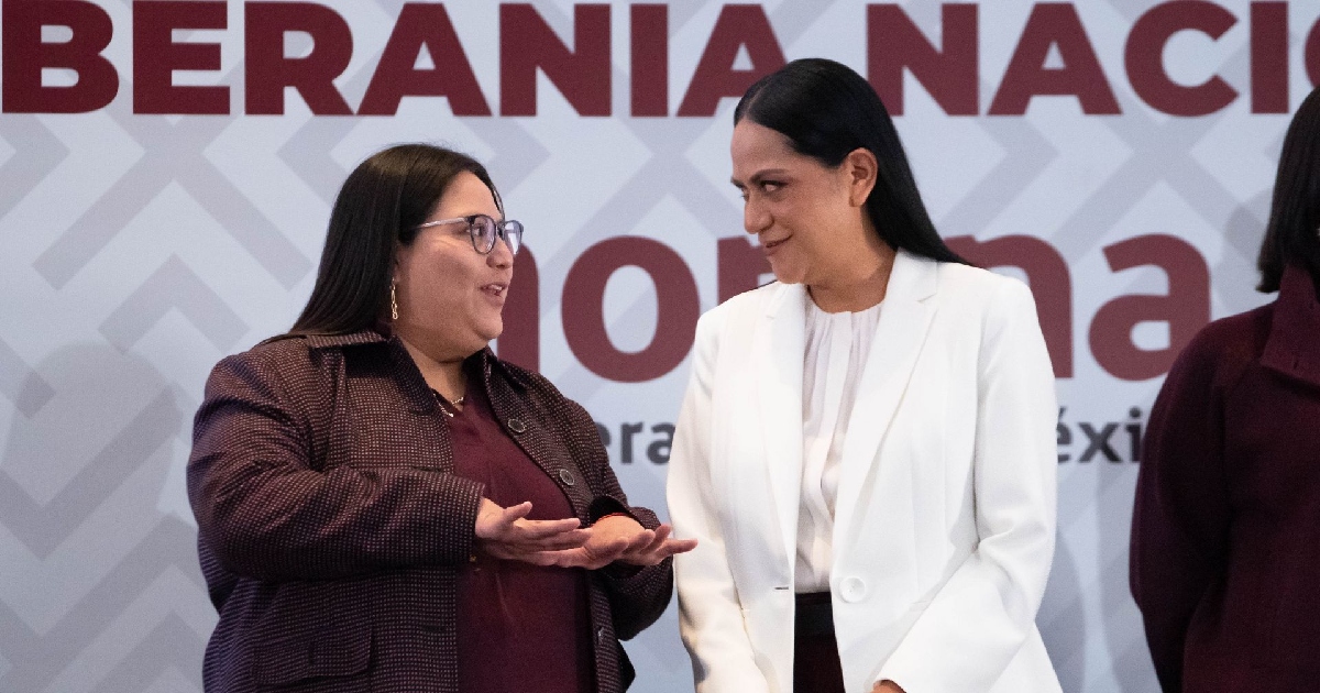 Citlalli Hernández Mora y Ariadna Montiel Reyes conversando amablemente durante un evento institucional o político. A la izquierda, Citlalli Hernández viste un abrigo café oscuro sobre blusa guinda y anteojos, gesticulando con ambas manos abiertas mientras habla. A la derecha, la secretaria de Bienestar, Ariadna Montiel, viste un saco blanco sobre blusa del mismo tono, sonriendo ligeramente mientras la escucha atentamente. Al fondo se observa una mampara oficial con patrones geométricos y letras en tonos guinda con la leyenda «SOBERANÍA NACIONAL».