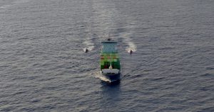 Vista aérea en plano general de un buque portacontenedores navegando en mar abierto. La embarcación lleva contenedores de carga apilados en su cubierta y avanza custodiada por dos embarcaciones de menor tamaño (patrulleras o lanchas de interceptación) que navegan a sus flancos, dejando estelas de agua en la superficie marina.
