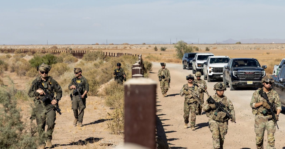 operativo militar y de seguridad fronteriza en una zona desértica. En primer plano y alineados a lo largo de un camino de tierra junto al muro y bolardos fronterizos, patrullan a pie varios soldados armados luciendo uniformes tácticos camuflados, cascos y chalecos antibalas. A la izquierda, agentes con uniformes oscuros verdes de tipo táctico avanzan paralelamente al muro. Detrás del contingente de infantería se observa una hilera de camionetas oficiales (pickup y SUV) escoltando el patrullaje bajo un cielo despejado.