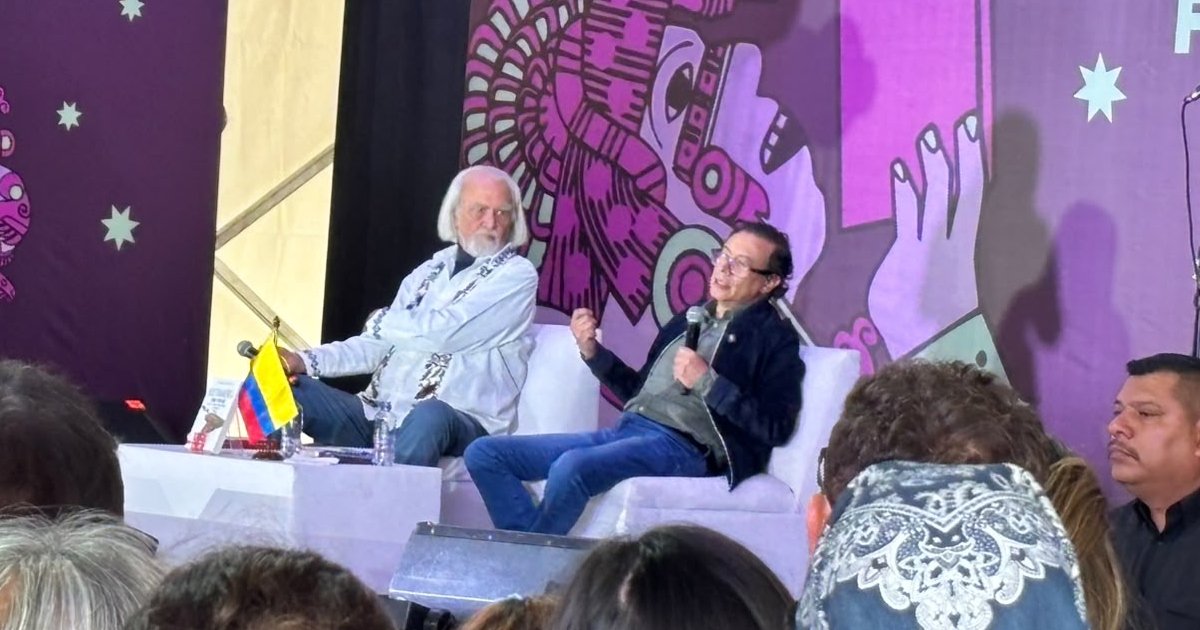 Gustavo Petro participando en un conversatorio en la Feria Internacional del Libro (FIL) del Zócalo de la Ciudad de México. Sentado en un sillón blanco junto al intelectual y escritor Armando Bartra, Petro viste una chaqueta azul marino sobre una camisa gris y jeans azules