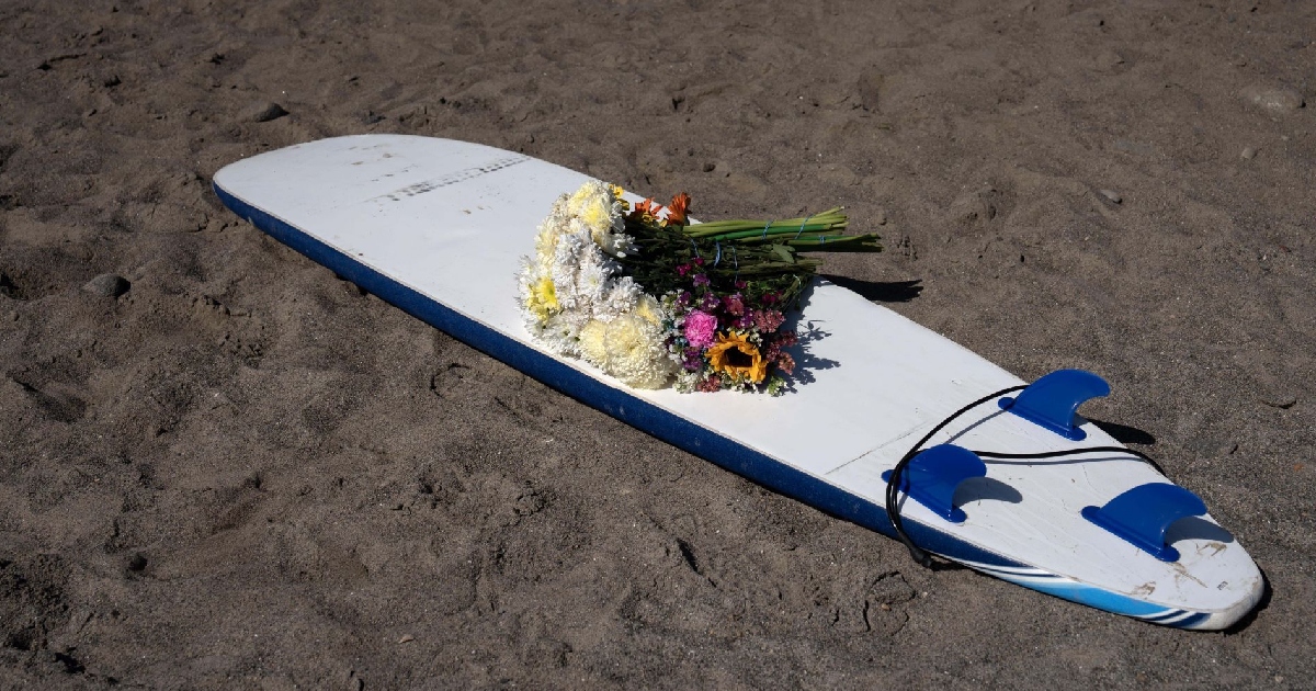 Plano medio cenital en ángulo picado sobre una tabla de surf blanca con bordes y quillas en azul claro reposada en la arena de una playa. Sobre el centro de la tabla descansa un arreglo floral a manera de homenaje y memoria, compuesto por crisantemos blancos, claveles rosas, girasoles y pequeñas flores silvestres multicolores. La luz natural resalta la textura de la arena y las sombras proyectadas alrededor del tributo memorial.