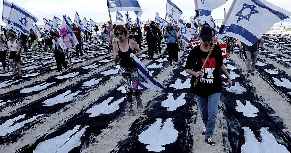 Una multitud de manifestantes ondea banderas israelíes mientras camina sobre la arena entre largas lonas o lienzos negros extendidos sobre el suelo. Dichas lonas tienen pintadas siluetas blancas de cuerpos humanos, simbolizando a las víctimas o rehenes del conflicto armado
