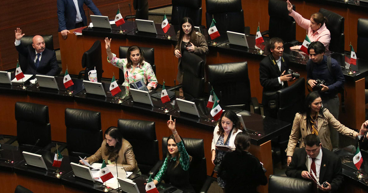 leno del Senado de la República Mexicana durante una sesión de votación. Se observa a varios senadores y senadoras sentados en sus escaños de piel negra con pequeñas banderas de México sobre sus escritorios; algunos de ellos levantan la mano para emitir su voto a favor o en contra de un dictamen. En los pasillos transita personal técnico y fotógrafos de prensa capturando la jornada legislativa.
